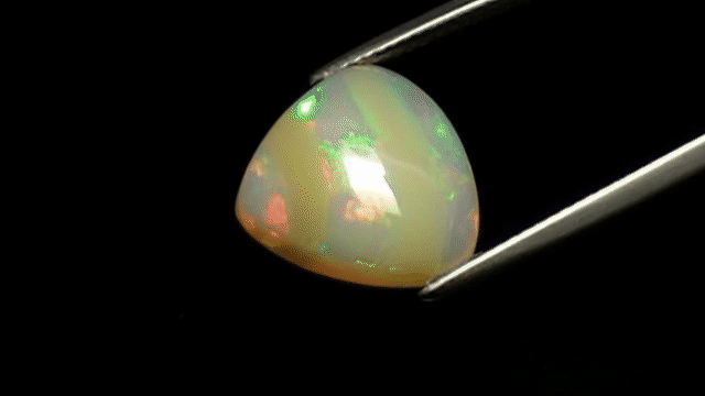 A pale opal turning between tweezers as green, red, gold, and blue flashes move across it.