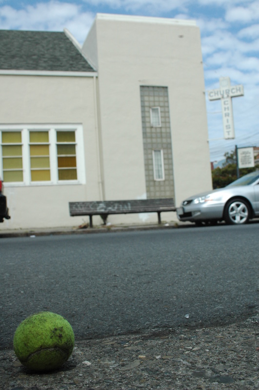 A stray tennis ball on the asphalt, with a white building and parked car beyond it.